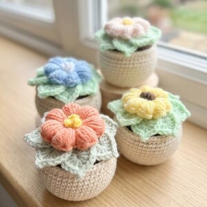May include: Four handmade crochet flower pots in pastel colors. Each pot features a different colored flower: pink, blue, peach, and yellow. The flowers sit atop green crocheted leaves, and the pots are a neutral beige color.
