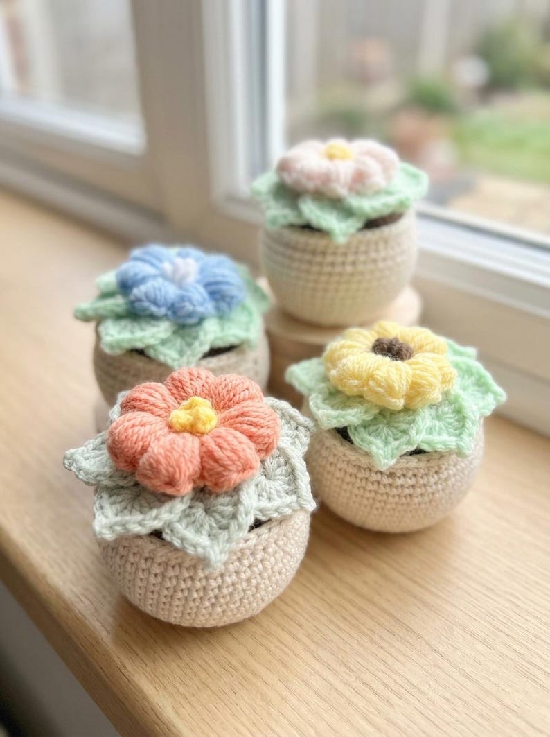 May include: Four handmade crochet flower pots in pastel colors. Each pot features a different colored flower: pink, blue, peach, and yellow. The flowers sit atop green crocheted leaves, and the pots are a neutral beige color.