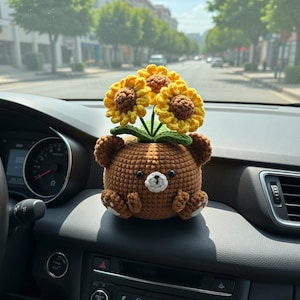 May include: A crocheted brown bear with yellow sunflowers on its head, sitting on a car dashboard. The bear has a cute face and small paws. The sunflowers have yellow petals and brown centers.