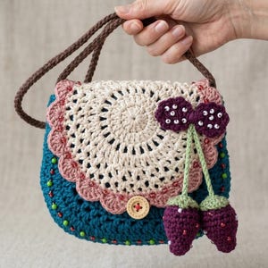 Könnte beinhalten: Handgefertigte Häkeltasche in Türkis, Creme und Rosa, verziert mit einem Holzknopf und Perlenakzenten. Die Tasche hat ein kreisförmiges cremefarbenes Design auf der Vorderseite und ist mit lila erdbeerförmigen Applikationen verziert.