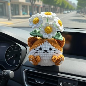 Op de afbeelding: Gehaakte katvormige auto-ventilatieopening decoratie met een boeket witte en gele madeliefjes. De kat is wit en oranje, met zwarte geborduurde gezichtskenmerken. De bloemen hebben groene bladeren. De decoratie is op een autodashboard geplaatst.