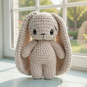 Könnte beinhalten: Ein beigefarbener Häkelhase mit langen, schlaffen Ohren und schwarzen Knopfaugen. Das handgefertigte Stofftier hat ein strukturiertes Aussehen und steht aufrecht. Der Hase steht vor einem Fenster, was auf ein weiches, natürliches Licht hindeutet.