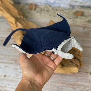 Könnte beinhalten: Handgefertigter, marineblauer Häkelrochen mit grauen Akzenten. Das Spielzeug hat schwarze Knopfaugen und wird auf einer Holzoberfläche präsentiert. Der Rochen ist ein weiches, plüschiges Spielzeug.