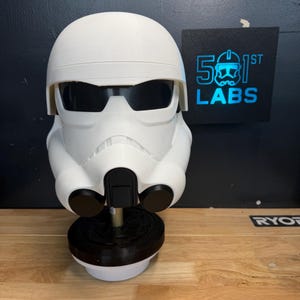 May include: A white Stormtrooper helmet with black lenses, displayed on a black and white stand. The helmet is a replica from the Star Wars franchise. A black sign with the text "501st LABS" is in the background.