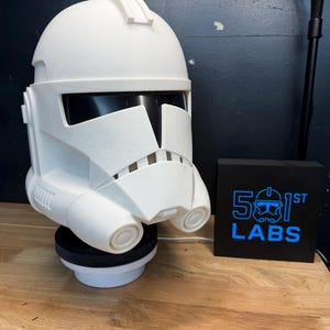 May include: A white, 3D-printed Clone Trooper helmet, resting on a black and white stand. The helmet features a detailed design with black visor and ventilation details. A black box with the text "501st LABS" in blue is to the right.
