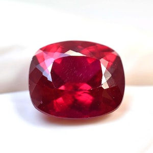May include: A faceted, cushion-cut ruby gemstone. The gem is a deep, saturated red color with multiple facets that reflect light. The shape is square with rounded corners, and the overall appearance is polished and brilliant.
