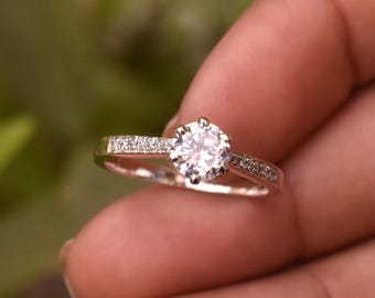 May include: A silver engagement ring with a large, round, clear gemstone as the centerpiece. The band is also adorned with smaller, clear gemstones. The ring is held up against a blurred green background.