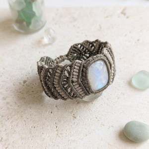 May include: A macrame bracelet featuring a central rectangular moonstone with a blue iridescent sheen. The band is crafted from taupe-colored cord in a repeating chevron pattern, creating a textured, woven design that encircles the wrist.