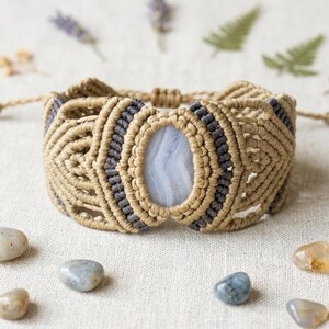 May include: A macrame bracelet featuring a central oval light blue banded stone. The bracelet is crafted from tan and dark gray cord, arranged in intricate knotted patterns that surround the stone. The design includes adjustable cord ties.