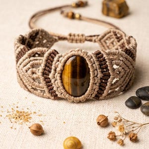 May include: A macrame bracelet featuring a central oval tiger's eye gemstone with a faceted surface. The band is crafted from tan and dark brown cord, woven into intricate geometric patterns with adjustable pull strings and small bead accents.