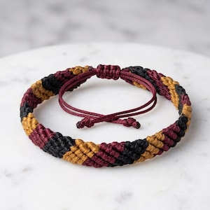 May include: A woven cord bracelet featuring a repeating diagonal pattern of black, mustard yellow, and deep burgundy threads. The bracelet includes an adjustable sliding knot closure with two thin burgundy cords ending in small knotted tips.