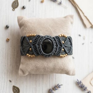 May include: A dark grey macrame bracelet featuring a central oval black stone, intricate knotted cord patterns, and small metallic gold-toned beads arranged symmetrically along the band, displayed on a soft beige square fabric cushion.