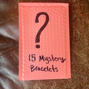 May include: A pink bubble mailer with a large black question mark and the handwritten text "15 Mystery Bracelets". The mailer is on a brown leather-like surface.