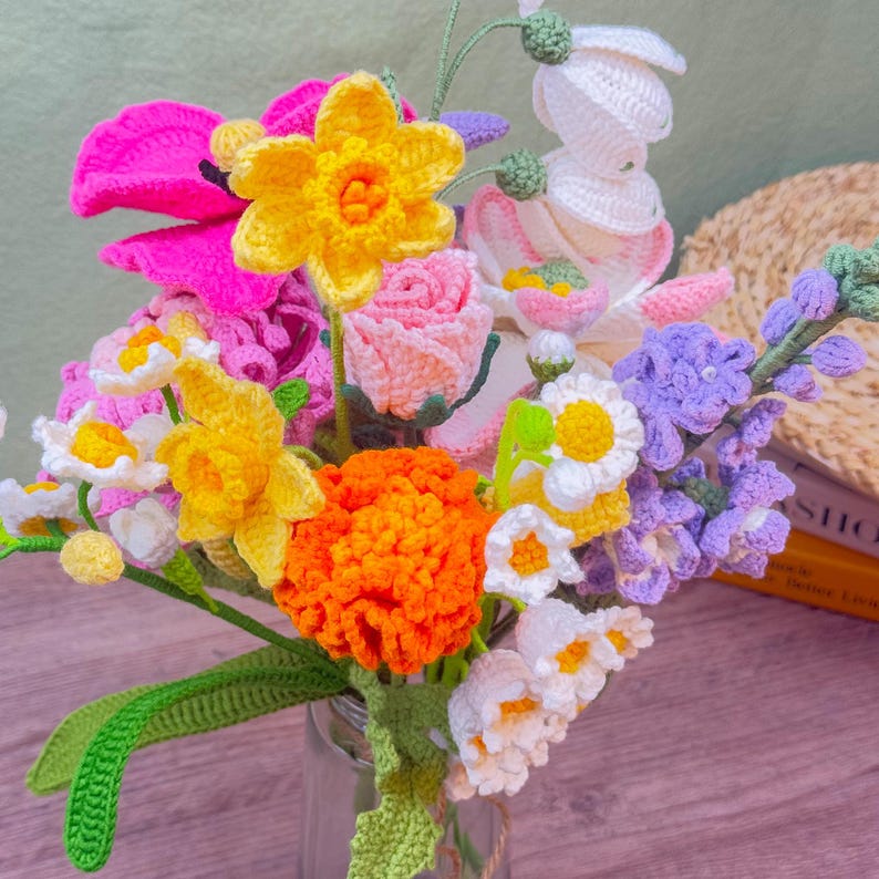 May include: A colorful bouquet of crocheted flowers in a glass vase. The arrangement includes yellow daffodils, pink poppies, orange marigolds, and white daisies. The handmade flowers have green stems and leaves, creating a vibrant and cheerful display.