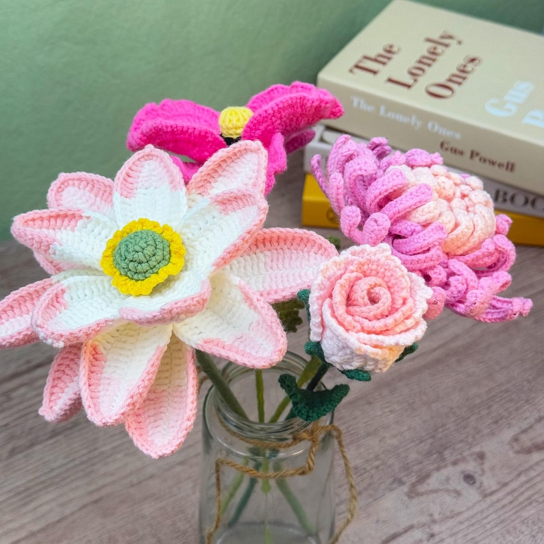 May include: A bouquet of handmade crochet flowers in a glass jar. The arrangement includes a large pink and white flower, a bright pink flower, a pink chrysanthemum, and a peach-colored rose. The flowers are displayed in a clear glass jar.