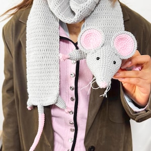 May include: A gray crocheted scarf with a mouse design. The mouse has pink ears, a pink nose, and black eyes. The scarf is wrapped around a person wearing a pink shirt and a brown jacket.