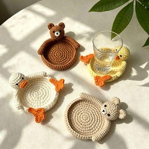 Könnte beinhalten: Vier handgefertigte Häkeluntersetzer mit Tiermotiven. Zwei Untersetzer sind als braune und beige Bären mit runden Ohren gestaltet. Zwei Untersetzer sind als weiße und gelbe Enten mit orangefarbenen Schnäbeln und Schwimmfüßen geformt.