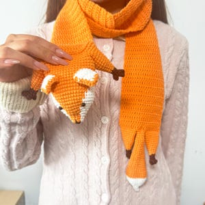 May include: A crocheted orange fox scarf with white and brown accents. The scarf features a fox head at one end and a tail design at the other. The fox's face has black eyes and a white muzzle.