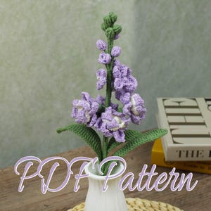 Può includere: Una composizione floreale all'uncinetto fatta a mano in un vaso bianco. La composizione presenta un lungo stelo con fiori viola e foglie verdi. Il testo "PDF Pattern" è sovrapposto all'immagine.