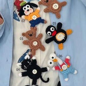 May include: A collection of six crocheted animal appliques featuring a dog, bears, a penguin, a cat, and a duck, each with a central white button. The figures are labeled with numbers 02, 03, 04, 05, and 06, showcasing various colors including black, brown, yellow, blue, red, and white yarn.