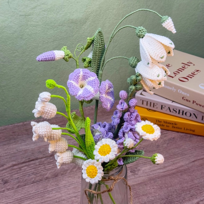May include: A handcrafted crochet bouquet of various flowers in a glass jar. The bouquet includes white bell-shaped flowers, purple morning glories, daisies, and other blooms. The flowers are arranged in a clear glass jar tied with twine.