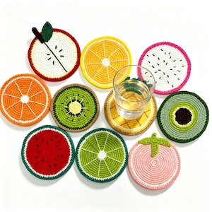 Op de afbeelding: Een verzameling van tien ronde, gehaakte onderzetters die lijken op verschillende soorten gesneden fruit, waaronder watermeloen, sinaasappel, kiwi, limoen, appel en drakenfruit, met een helder glazen beker op een onderzetter met ananaspatroon.