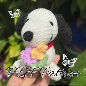 May include: A crocheted white dog with black ears and a black nose, wearing a red collar and a colorful flower arrangement. The text "PDF Pattern" is displayed at the bottom. Two butterfly graphics are in the background.