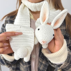 May include: A white crochet scarf and matching bunny-shaped hand warmers. The bunny has pink inner ears and a pink nose. The hand warmers have a bunny tail design. The scarf is a simple, open weave.