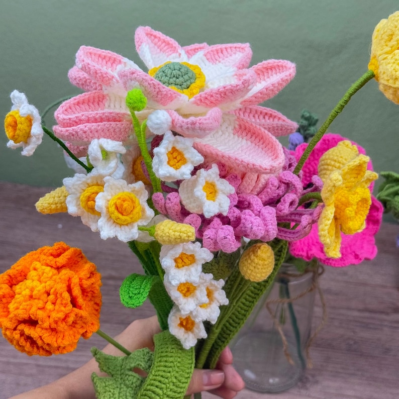 May include: A colorful bouquet of crocheted flowers, including pink lotus blossoms, yellow daffodils, and orange marigolds. The handmade arrangement features various flower types with green stems and leaves, creating a vibrant and textured display.