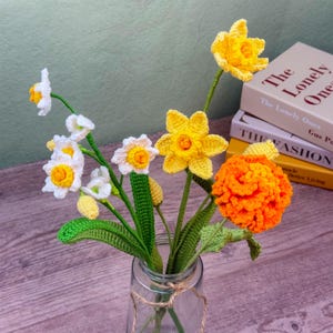 May include: A bouquet of handmade crochet flowers in a clear glass vase. The flowers include white and yellow daffodils, and an orange marigold. The vase is tied with twine, and the background includes books.