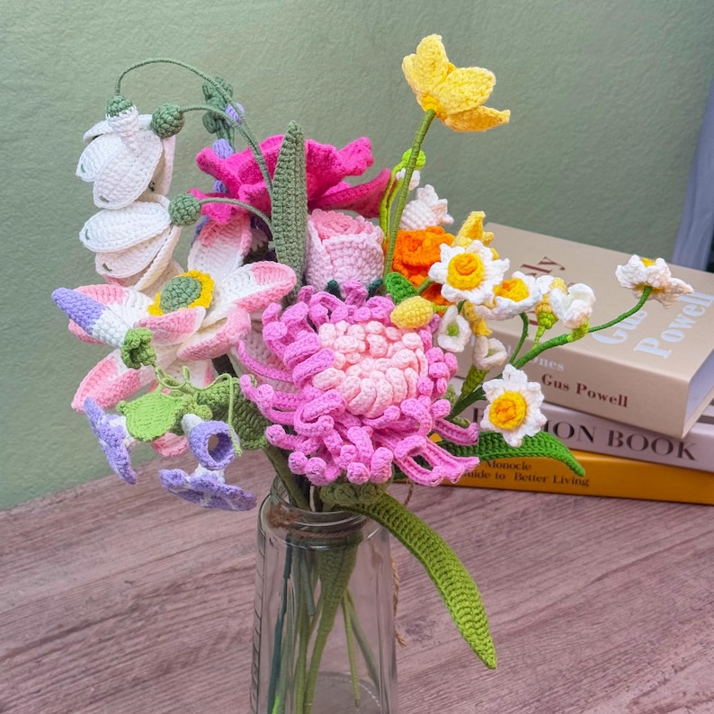 May include: A colorful bouquet of crocheted flowers in a clear glass vase. The arrangement includes pink, yellow, white, and purple flowers with green stems and leaves. The flowers are intricately crafted and appear handmade.