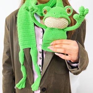Può includere: Una sciarpa a forma di rana all'uncinetto verde brillante con bocca beige e occhi marroni. La sciarpa presenta una testa e zampe di rana, creando un design stravagante. La sciarpa è realizzata in filato morbido.