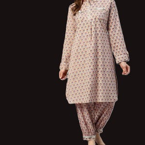 Könnte beinhalten: Beige zweiteiliges Outfit mit floralem Muster. Das langärmlige Oberteil hat einen Kragen und zwei Brusttaschen. Die passende Hose ist locker geschnitten mit gerafften Knöcheln. Weiße Paspeln akzentuieren die Manschetten und Taschen.