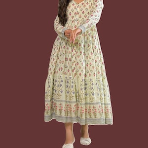 May include: A long, flowing dress with a light green background and a floral pattern in red, blue, and white. The dress has long sleeves and a tiered skirt. The dress is paired with white shoes.