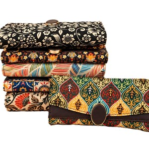 May include: A stack of five rectangular fabric wallets featuring diverse patterns including black and white floral, colorful geometric, and leaf motifs. Each wallet has a flap closure with a central oval or round button accent, and one wallet is placed horizontally in front of the stack.
