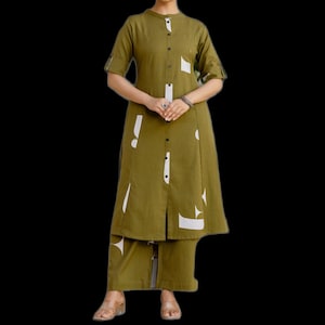 Op de afbeelding: Olijfgroene tweedelige outfit met witte geometrische accenten. De outfit bestaat uit een tuniek met knopen en korte mouwen en een bijpassende broek met wijde pijpen. De tuniek heeft een mandarijnkraag en witte geometrische vormen.