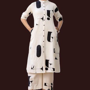 May include: A cream-coloured, two-piece outfit comprising a long-sleeved tunic and wide-leg trousers. The tunic has a mandarin collar and a button-down front. Bold black abstract shapes are printed across the outfit. The outfit has pockets on the tunic.