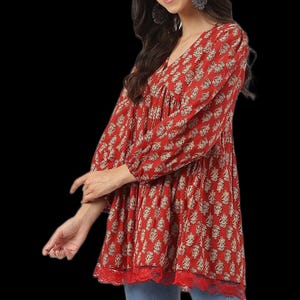Puede incluir: Una túnica roja con cuello en V y mangas largas. La parte superior presenta un estampado floral beige y un ribete de encaje en el dobladillo. La modelo lleva vaqueros azules.