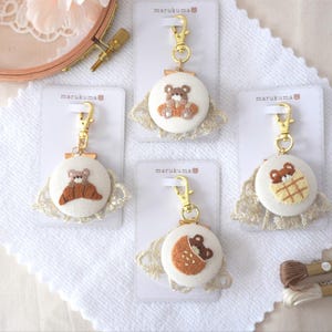 Könnte beinhalten: Vier handgefertigte Schlüsselanhänger mit gestickten Teddybären-Designs. Jeder Schlüsselanhänger hat eine runde, cremefarbene Stoffbasis mit einem goldfarbenen Verschluss und einer dekorativen Spitzentrimmung. Die Teddybären sind in verschiedenen Posen und Farben.