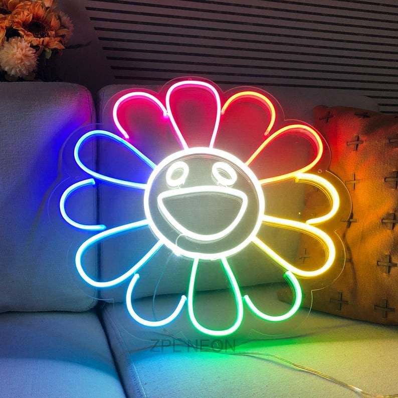 Pu&ograve; includere: Un'insegna al neon a forma di fiore sorridente con un gradiente arcobaleno. I petali passano dal rosso al giallo, al verde e al blu. Il viso e il contorno sono bianchi. L'insegna &egrave; su un divano di colore chiaro.