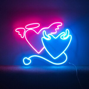 Pu&ograve; includere: Insegna al neon raffigurante due forme di cuore. Uno &egrave; rosa con ali d'angelo e un alone, l'altro &egrave; blu con corna di diavolo e una coda. L'insegna &egrave; illuminata da luci al neon rosa e blu brillante, creando un contrasto sorprendente.