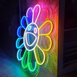Pu&ograve; includere: Un'insegna al neon a forma di fiore sorridente con una combinazione di colori arcobaleno. L'insegna ha un contorno bianco ed &egrave; montata su una superficie di legno. I colori al neon includono blu, verde, giallo, arancione e rosa.