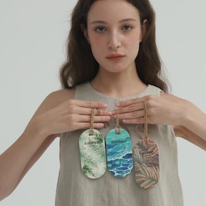 May include: Three wooden tags with natural designs and the word "EUOPHO" printed on them. The tags are oval-shaped and suspended by twine. The designs include ferns, ocean waves, and palm leaves. The tags are held in front of a person wearing a neutral-colored top.