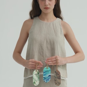 May include: A woman holds three oval-shaped air fresheners with tropical designs. The air fresheners have a string for hanging. The designs include palm leaves, ocean waves, and other tropical foliage. The woman is wearing a linen top and skirt.