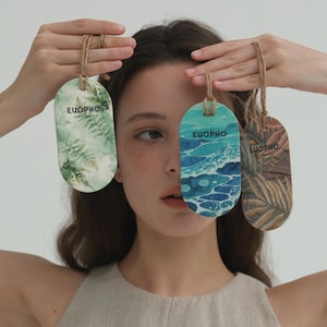 May include: Three oval-shaped tags with rope hangers are held up. Each tag features a different nature-themed design: ferns, ocean waves, and palm leaves. The word "EUOPHO" is printed on each tag. The tags are in shades of green, blue, and brown.