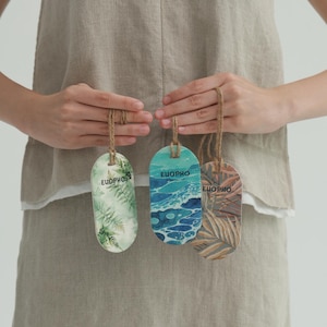May include: Three oval-shaped air fresheners with jute twine hangers. The designs include fern leaves, ocean waves, and palm fronds. Each has the word "EUOPHO" printed on it. The air fresheners are held against a neutral-colored linen garment.