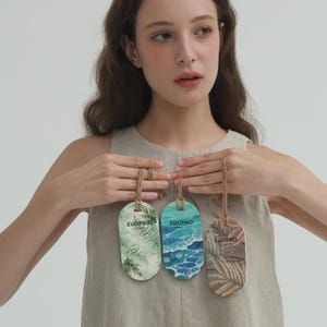 Könnte beinhalten: Drei dekorative Holzanhänger mit Naturmotiven und dem Aufdruck "EUOPHO". Die Anhänger werden von einer Person gehalten. Die Anhänger zeigen Bilder von Blättern, Meereswellen und Palmenwedeln. Jeder Anhänger ist mit einem Naturfaden befestigt.