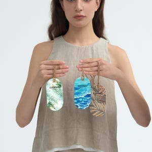 以下が含まれることがあります： 自然をテーマにしたデザインの、3つの楕円形の装飾用芳香剤が表示されています。それぞれに吊り下げ用の紐が付いています。デザインには、シダ、海の波、ヤシの葉が含まれています。「EUOPHO」の文字がそれぞれに印刷されています。