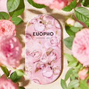 以下が含まれることがあります： ピンク色の楕円形のタグ。バラの花柄のデザインと「EUOPHO Encounter Love」の文字が特徴です。タグはピンク色のバラと緑の葉に囲まれ、ロマンチックでフローラルなテーマを演出しています。