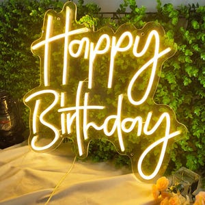 May include: A neon sign with the words "Happy Birthday" in a cursive font. The sign is illuminated with a warm, yellow glow and is set against a backdrop of green foliage. A clear plastic border surrounds the text.